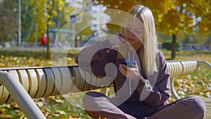 A blonde teenager girl sits on a bench in an autumn park, strewn with yellow leaves and looks at the phone.