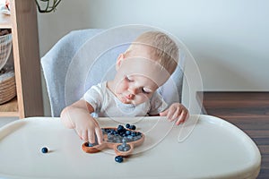 Blonde toddler boy eating Yummy blueberries with wooden plates on highchair close-up and copy space