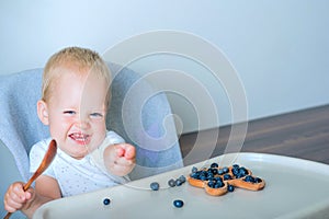 Blonde toddler boy eating Yummy blueberries wooden spoon on highchair close-up and copy space