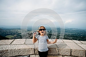 Blonde toddler girl in black sunglasses and striped t-shirt emotional smiling on top of Lovchen mountain in Montenegro.