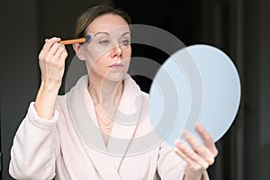 Blonde woman applying makeup with a brush and looking into a mirror