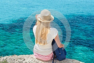 Blonde woman with backpack sitting on cliff above sea