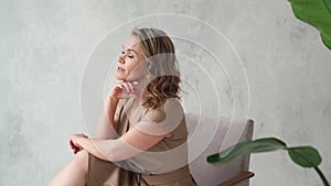 A blonde woman in a beige dress poses on a gray chair in a photo studio.