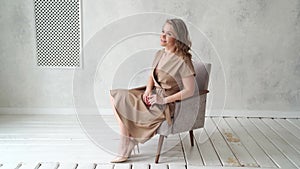 A blonde woman in a beige dress poses on a gray chair in a photo studio.