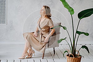 A blonde woman in a beige dress poses on a gray chair in a photo studio.