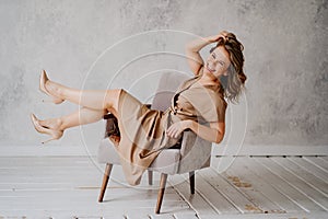 A blonde woman in a beige dress poses on a gray chair in a photo studio.