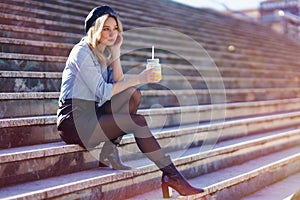 Blonde woman with beret, drinking a natural orange juice in a crystal glass, sitting on some steps.