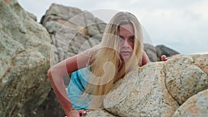 Blonde woman in blue dress crawls on rocks. Outdoor adventure, untamed spirit, natural landscape. Elegant, wild