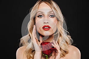 Blonde woman with bright makeup red lips posing isolated over black wall background holding rose flower