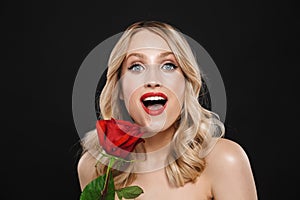 Blonde woman with bright makeup red lips posing isolated over black wall background holding rose flower
