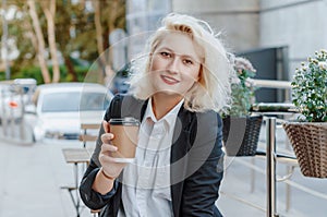 Blonde woman in a business suit sitting at a table in a street cafe, holding a cup of coffee in his hand