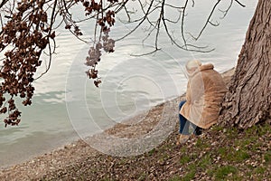 Blonde woman in coat sitting under the tree by the lake