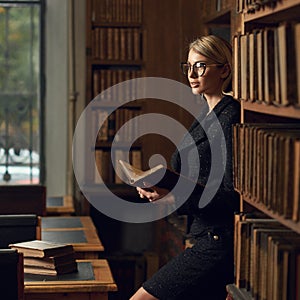 Blonde woman dressed in black tweed suit reading book in library