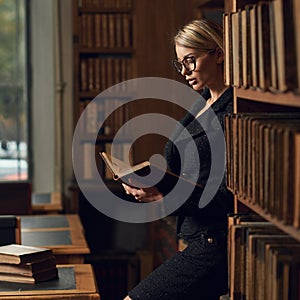 Blonde woman dressed in black tweed suit reading book in library