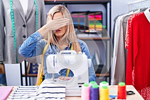 Blonde woman dressmaker designer using sew machine covering eyes with hand, looking serious and sad