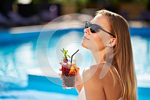 Blonde woman drinks refreshing cocktail sunbathing and sitting near swimming pool at tropical spa. Female in sunglasses