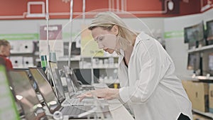 A blonde woman in an electronics hypermarket chooses a laptop. A lady in an electronics shop buys a computer