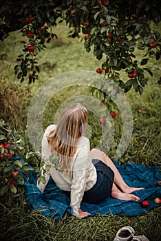 Blonde woman enjoying autumn day in the garden sitting on a blanket under apple tree full of tasty red apples