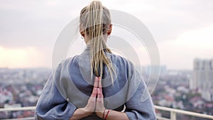 Blonde woman enjoys city view and folds her hands behind back in a namaste gesture