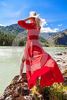 A blonde woman with fluttering hair and a red dress in the wind stands on the banks of the Katun River in the Altai mountains,