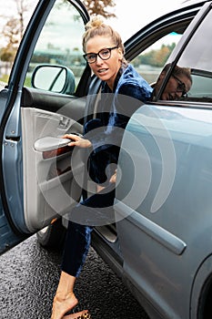 Blonde woman in glasses look back over her shoulder as she exits parked blue car in which she drove.