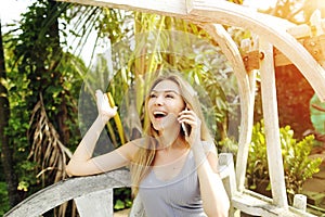 Blonde woman got great news by talking on smartphone for work on sunny day, background of sunshine green palms in