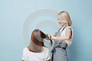 Blonde woman hairdresser straightening client`s hair with flat iron over blue