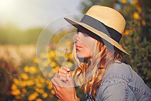 Blonde woman in hat blowing dandelion at summer