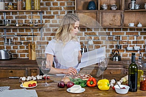 Blonde woman holding cookbook while preparing pizza in kitchen