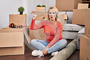 Blonde woman holding keys of new home sitting on the floor scared and amazed with open mouth for surprise, disbelief face