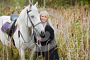 Blonde woman holds by bridle white horse among the