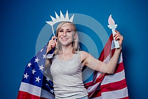 Blonde woman in jeans holding American flag with paper crown and torch Statue of liberty on a blue background in the