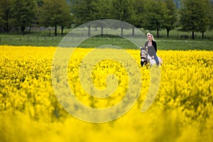 Laughing blonde woman sits on her horse in a blooming rapeseed field, houses and forest are out of focus in the background.