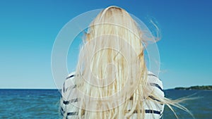 Blonde woman with long hair looks at the sea. Wind ruffles her hair