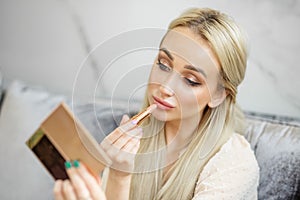 A blonde woman paints lips with beige lipstick. Small mirror in hand