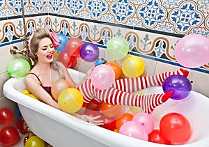 Blonde woman playing in her bath tube with bright colored balloons. Sensual girl with white red striped stockings