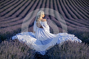 Blonde woman poses in lavender field at sunset. Happy woman in white dress holds lavender bouquet. Aromatherapy concept