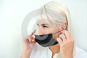 The blonde woman puts on a black protective mask on her face on a white background. Coronavirus protection.