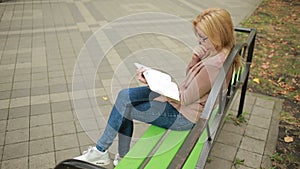 Blonde Woman Reading Book on Brench in Autumn Park.