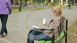 Blonde Woman Reading Book on Brench in Autumn Park. drinking hot coffee or tea