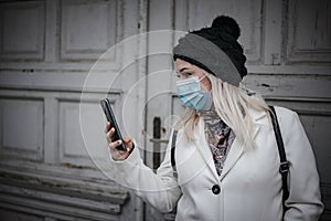 Blonde woman is reading a message while leaving house and wearing face mask