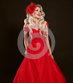 Blonde woman in red dress and elegant hat isolated on studio black background. Beautiful blond girk with long wavy hair and