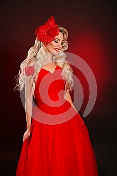 Blonde woman in red dress and elegant hat isolated on studio black background. Beautiful blond girk with long wavy hair and makeup
