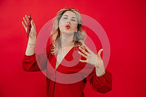 Blonde woman in red outfit blow and cool skin from hot pepper in hands, shaking arm and looking at camera in red studio