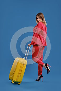 Blonde woman in red pantsuit, high black heels. She has raised her leg, posing back to the camera with yellow suitcase