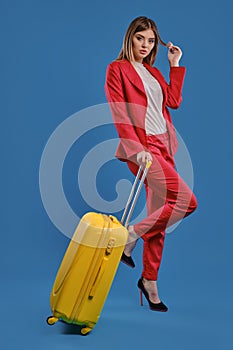 Blonde woman in red pantsuit, white blouse and high black heels. She is holding yellow suitcase, posing against blue