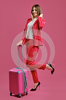 Blonde woman in red pantsuit, white blouse, high black heels. She smiling, posing with suitcase on pink background