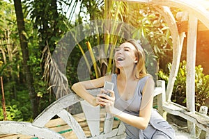 Blonde woman rejoice holding smartphone on sunny day, background of sunshine green palms in Thailand, Phuket travel