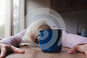 Blonde woman resting her head on a kitchen table cup of coffee in front of her