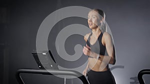 Blonde woman is running on treadmill in fitness club in dark hall, training her endurance, cardio workout